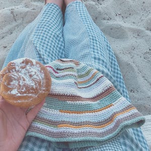 May include: A person wearing blue and white checkered pants sits on a sandy surface holding a pastry topped with powdered sugar. A multi-colored crocheted blanket is draped over their legs.