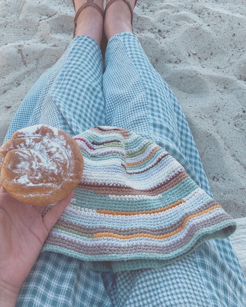 May include: A person wearing blue and white checkered pants sits on a sandy surface holding a pastry topped with powdered sugar. A multi-colored crocheted blanket is draped over their legs.