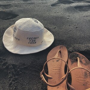 May include: A white crochet sun hat with the text "FREAKY FOR YAMR" on the front. The hat is sitting on black sand next to two brown flip-flops.
