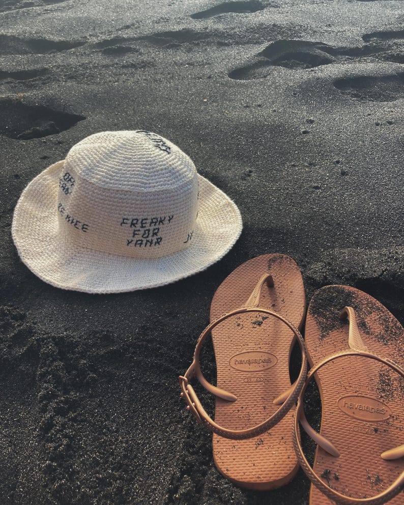 May include: A white crochet sun hat with the text "FREAKY FOR YAMR" on the front. The hat is sitting on black sand next to two brown flip-flops.
