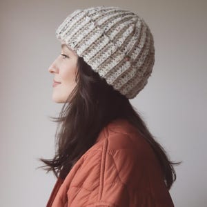 May include: A person wearing a beige crocheted beanie with a speckled pattern. The beanie is ribbed and has a slouchy fit. The person is wearing a rust-colored quilted jacket.