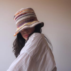May include: A person wearing a white shirt and a multi-colored crocheted bucket hat. The hat has stripes of brown, orange, yellow, and purple.