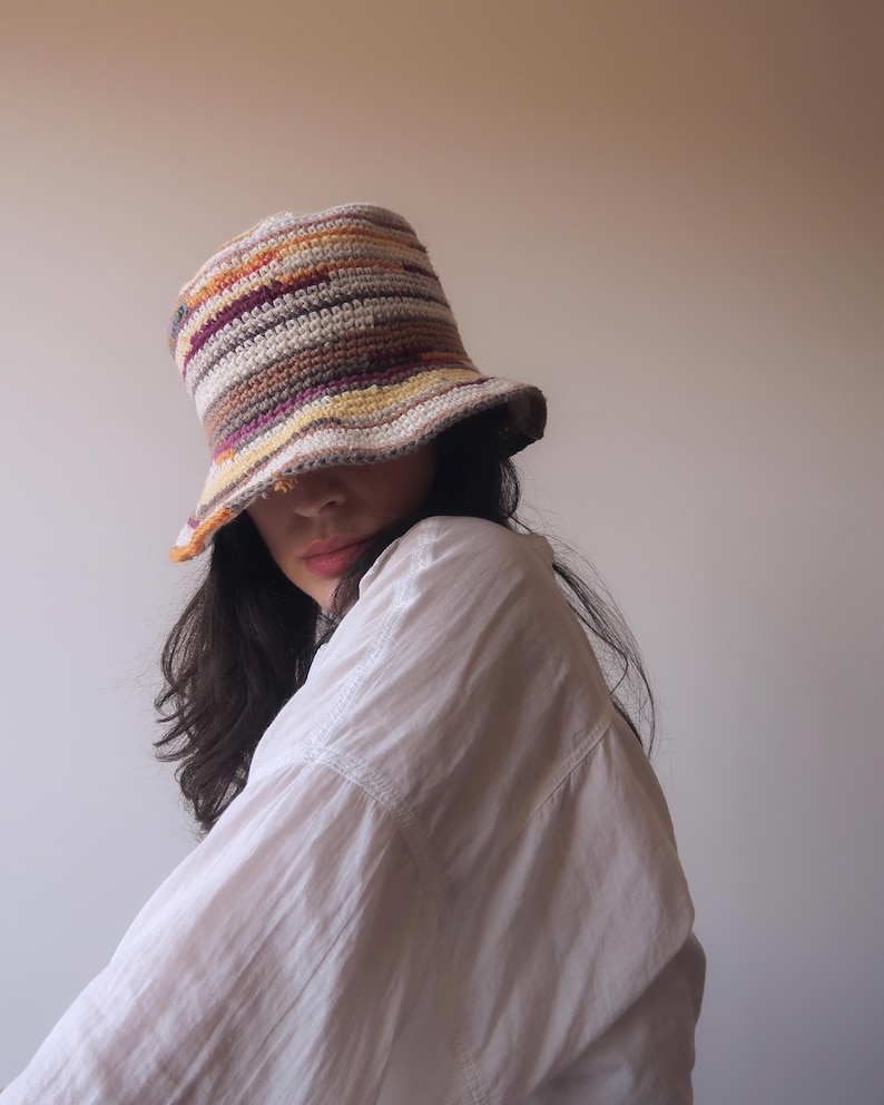 May include: A person wearing a white shirt and a multi-colored crocheted bucket hat. The hat has stripes of brown, orange, yellow, and purple.