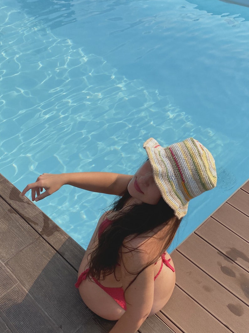 May include: A person wearing a striped crochet bucket hat and a red bikini top, sitting on the edge of a pool.
