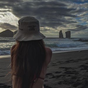 May include: A person wearing a white crochet hat with the text "FREAKY FOR YARN" sitting on a black sand beach looking out at the ocean. The ocean is choppy with white waves. There are two rock formations in the distance.
