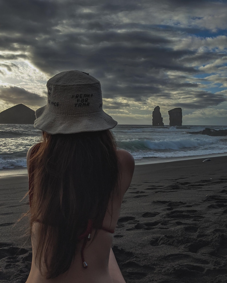 May include: A person wearing a white crochet hat with the text "FREAKY FOR YARN" sitting on a black sand beach looking out at the ocean. The ocean is choppy with white waves. There are two rock formations in the distance.