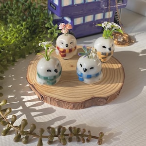 Op de afbeelding: Vier kleine keramische uilbeeldjes met verschillende gekleurde sjaals, elk met een klein boeket witte bloemen erop. De uilen zitten op een houten schijf.