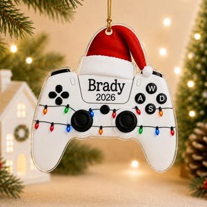 May include: A white video game controller ornament featuring a red Santa hat on top, a string of colorful Christmas lights, and the text "Brady 2026" printed in black. The controller includes black analog sticks and buttons labeled W, A, S, and D.