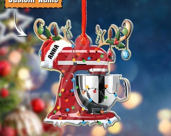 May include: A red and silver Christmas ornament shaped like a stand mixer, adorned with colorful lights and a Santa hat. The ornament has a custom name and is hanging from a red string. The background is blurred with Christmas tree and lights.
