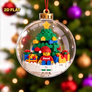 May include: A clear spherical Christmas ornament containing a buildable brick-style green tree with yellow and blue accents, a yellow star topper, and a small figure wearing a red hat and shirt with the name David printed in white.