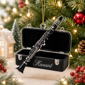 May include: A black clarinet ornament with silver-toned keys resting diagonally across an open, rectangular black case. The case features a clear acrylic lid and the name Hanant printed in white script on the front exterior surface.