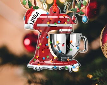 May include: A red Christmas ornament shaped like a stand mixer, decorated with colorful lights. The ornament has a Santa hat with the name "Linda" on it and reindeer antlers. A silver mixing bowl is attached to the mixer.