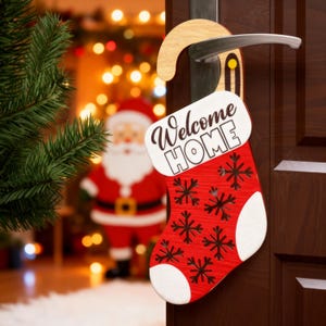 May include: A red and white Christmas stocking-shaped door hanger with the words "Welcome HOME" in black script. The stocking features snowflake cutouts and hangs from a wooden hook on a brown door. A Christmas tree and Santa Claus are in the background.