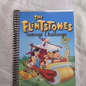 May include: A spiral-bound notebook featuring a colorful cover with the title "The Flintstones Savings Challenge." The cover art depicts the Flintstones family in their iconic car. The text "Saving $1000+" and "Cajun Girl Creates" are also visible.