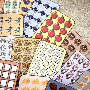 K&ouml;nnte beinhalten: Eine Sammlung farbenfroher, rechteckiger Aufkleber mit verschiedenen Designs. Die Aufkleber zeigen Bilder von &Auml;pfeln, W&uuml;rfeln, Zeichentrickfiguren, Pfotenabdr&uuml;cken und anderen Mustern. Die Aufkleber sind f&auml;cherf&ouml;rmig angeordnet.