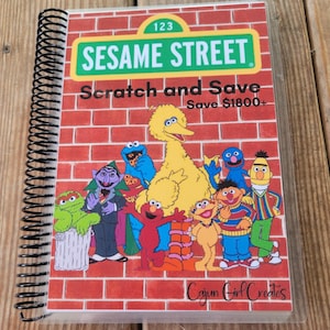 May include: A Sesame Street themed scratch and save book with a red brick background. The book features Big Bird, Cookie Monster, Elmo, and other Sesame Street characters. The book cover says "Sesame Street Scratch and Save Save $1800+"