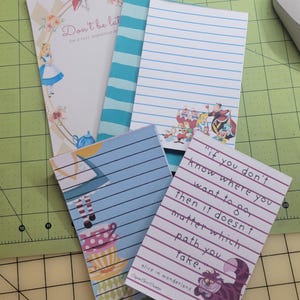 Op de afbeelding: Een verzameling notitieblokken met het thema Alice in Wonderland. Eén notitieblok toont Alice, een ander heeft blauwe strepen en andere hebben citaten en illustraties van personages. De notitieblokken hebben gelinieerde pagina's en decoratieve randen.