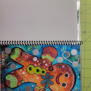 May include: A colorful gingerbread man illustration with a blue flower and green buttons. The illustration is surrounded by silver dots on a blue background. The text "Save $5.50" is in the top left corner.