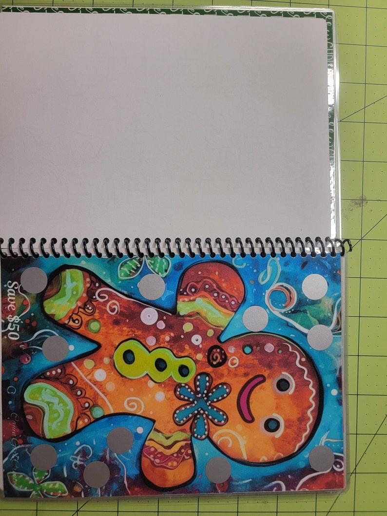 May include: A colorful gingerbread man illustration with a blue flower and green buttons. The illustration is surrounded by silver dots on a blue background. The text "Save $5.50" is in the top left corner.