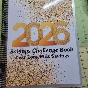 Puede incluir: Un planificador encuadernado en espiral con una cubierta blanca que presenta el año "2026" en purpurina dorada. El texto "Savings Challenge Book Year Long Plus Savings" está debajo. Salpicaduras doradas y marrones decoran la cubierta.