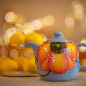 Peut inclure: Une théière en céramique bleu clair avec un grand motif floral coloré. La fleur est principalement orange et jaune avec des accents rouges. Un bocal en verre de citrons est derrière la théière, avec un arrière-plan flou de lumières chaudes.