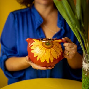 Peut inclure: Une théière en céramique rouge avec un motif de tournesol. La théière a un couvercle argenté et une poignée. La théière est tenue par une personne.