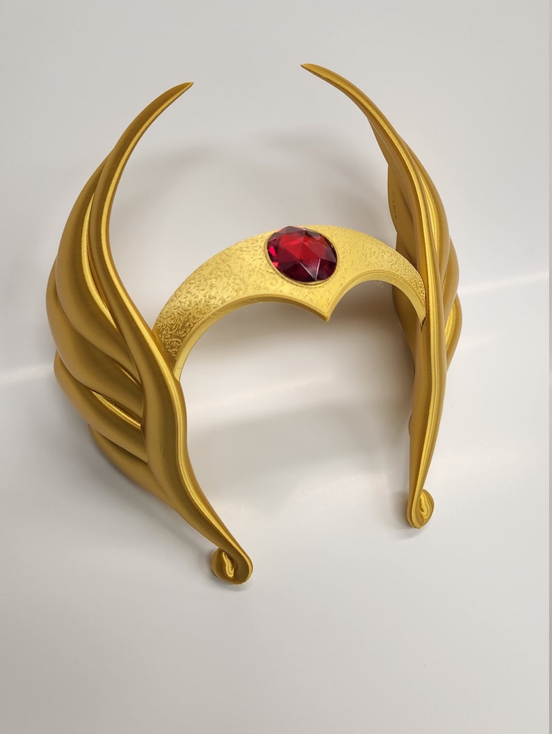 May include: A gold crown with a red jewel in the center. The crown has a wing-like design on each side.