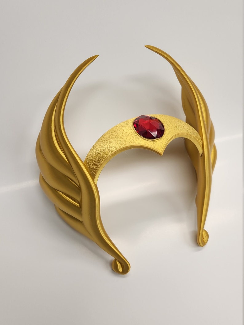 May include: A gold-colored crown with a red jewel in the center. The crown has a wing-like design on each side.
