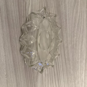 May include: A clear glass ashtray with a distinctive geometric pattern. The ashtray features a central oval area and is surrounded by diamond-shaped facets. The edges are scalloped, enhancing its decorative look. The ashtray is empty and rests on a light-coloured wooden surface.