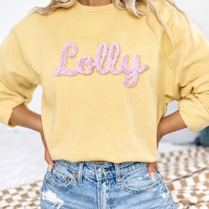 May include: A yellow sweatshirt with the name "Lolly" in pink, textured lettering. The sweatshirt is a solid color and has long sleeves. The person is wearing blue denim jeans.
