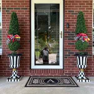 May include: Two topiary plants in black and white urns flank a white door. The urns have a black and white striped design with gold accents. Each topiary has a cone-shaped top and a round base, adorned with a colorful bow.