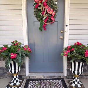 May include: A gray front door with a green wreath featuring a pink and green patterned ribbon. Two black and white striped urns hold pink flowers and green foliage. A rectangular doormat displays a central bow and the text WELCOME.