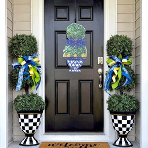 May include: A front door with a black finish and a decorative topiary design hanging on it. Two potted topiary trees flank the door, each adorned with blue and yellow ribbons. The pots have a black and white checkered pattern. A welcome mat is on the floor.