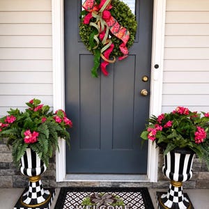 May include: A front door with a green wreath and a large pink and red bow. Two black and white striped planters with pink flowers flank the door. A welcome mat with a wreath design sits in front of the door.