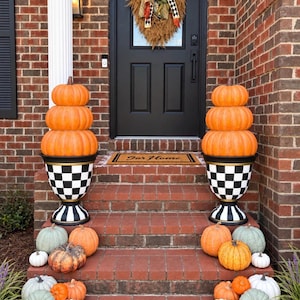 May include: Two black and white checkered urns each topped with a stack of three orange pumpkins. The steps are decorated with various pumpkins in shades of orange, white, and sage green. A black door features a wreath with ribbons and a mat reading Our Home.