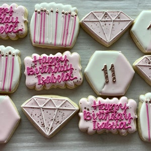May include: A collection of decorated sugar cookies. Some cookies are shaped like diamonds, hexagons, and rectangles. The cookies are decorated with pink frosting, sprinkles, and the words "Happy Birthday Isabella" in pink icing. Some cookies have the number "11".