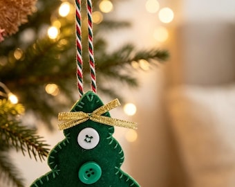 Può includere: Un addobbo natalizio a forma di albero in feltro verde con un fiocco in nastro dorato, tre bottoni decorativi di colore bianco, verde e a quadri rossi e bianchi, appeso tramite un cordoncino intrecciato nei colori rosso, bianco e verde.