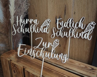 Może przedstawiać: Trzy drewniane tabliczki z tekstem "Hurra Schulkind", "Endlich Schulkind" i "Zur Einschulung" z małą grafiką czapki imprezowej obok każdego zdania.