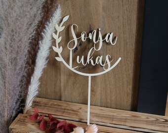 Może przedstawiać: Drewniany topper na tort z okrągłym wzorem i imionami "Sonja" i "Lukas" napisanymi kursywą. Topper stoi na drewnianej podstawce i jest otoczony suszonymi kwiatami.