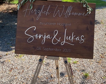 Może przedstawiać: Drewniany znak z napisem "Herzlich Willkommen zu unserer Hochzeit Sonja & Lukas 08 September 2023" w białym kolorze. Znak jest ustawiony na drewnianym sztalugach.