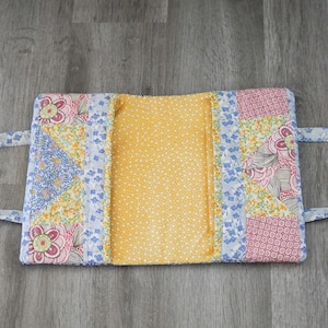 May include: A quilted book cover with a yellow interior and floral patchwork exterior. The exterior features pink, blue, and yellow floral patterns. It has two fabric handles for carrying.