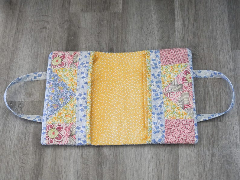May include: A quilted book cover with a yellow interior and floral patchwork exterior. The exterior features pink, blue, and yellow floral patterns. It has two fabric handles for carrying.