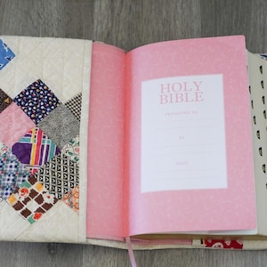 May include: Open Bible with a pink cover and pages, featuring the text "Holy Bible." The cover is a patchwork design with various fabric patterns and colors, including blue, pink, and brown. A pink ribbon bookmark is visible.