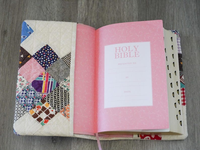 May include: Open Bible with a pink cover and pages, featuring the text "Holy Bible." The cover is a patchwork design with various fabric patterns and colors, including blue, pink, and brown. A pink ribbon bookmark is visible.