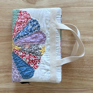 May include: A handmade fabric book cover with a colorful, fan-shaped design. The cover features a variety of patterned fabrics in red, blue, purple, and yellow. It has a white handle and a white fabric spine.