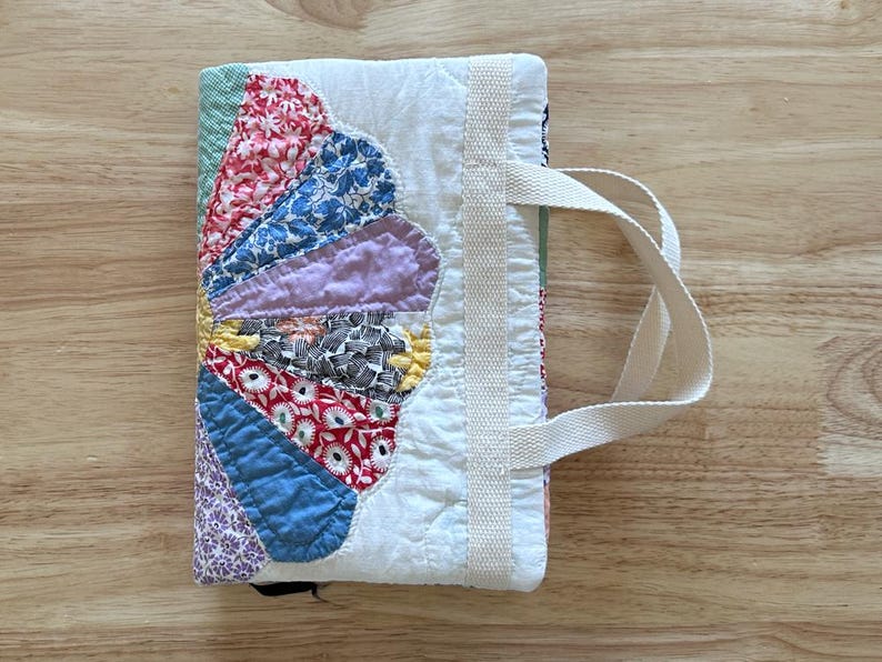 May include: A handmade fabric book cover with a colorful, fan-shaped design. The cover features a variety of patterned fabrics in red, blue, purple, and yellow. It has a white handle and a white fabric spine.