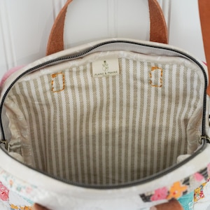 May include: Open view of a cream-colored handbag with a floral patchwork design. The bag has a brown leather handle and strap, and a striped interior. The interior has the text "PLAINS & PRAIRIE".