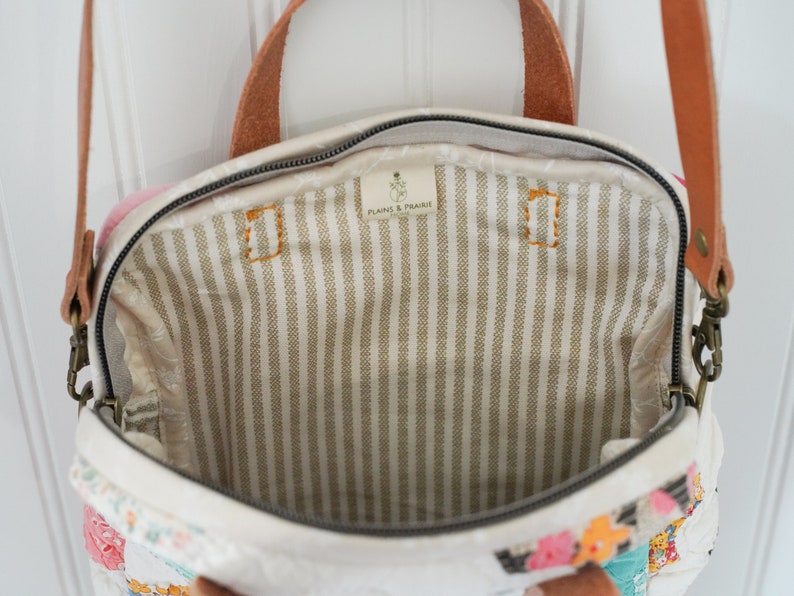 May include: Open view of a cream-colored handbag with a floral patchwork design. The bag has a brown leather handle and strap, and a striped interior. The interior has the text "PLAINS & PRAIRIE".