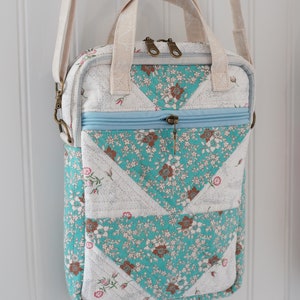 May include: A quilted crossbody bag with a floral pattern in teal, brown, and pink. The bag has a light blue zipper and a cream-colored strap and handles. The bag is rectangular with rounded corners.