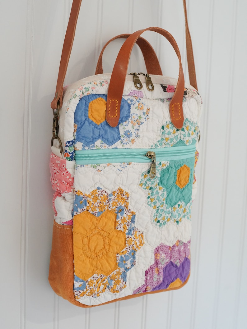 May include: A quilted crossbody bag with a patchwork design featuring floral patterns in blue, yellow, and purple. The bag has a light blue zipper, brown leather handles, and a brown leather shoulder strap. The bottom of the bag is a solid orange color.